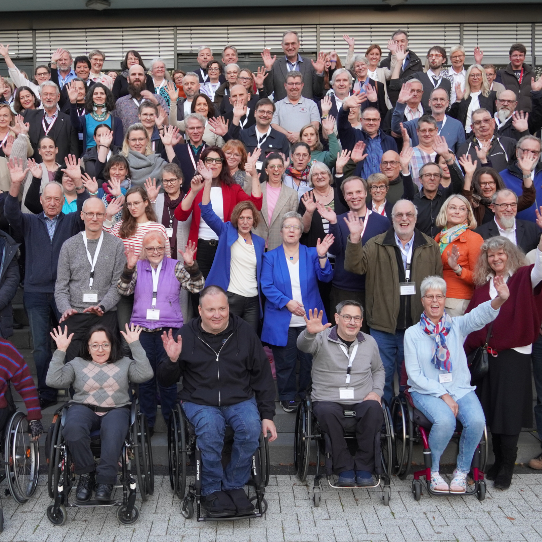 Gruppenbild_MV_2025 Große Gruppe von Menschen, darunter mehrere Personen im Rollstuhl, posiert vor einem mehrstöckigen Gebäude auf einer Treppe und winkt in die Kamera.