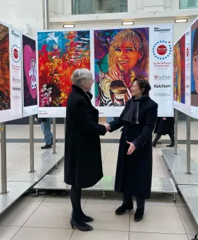 2 Frauen in schwarzem Mantel stehen vor einer Stehle mit buntem Bild und schütteln sich die Hand. Es sind Humangenetikerin Prof. Evelin Schröck und Eva Luise Köhler in Mission für die ACHSE. 