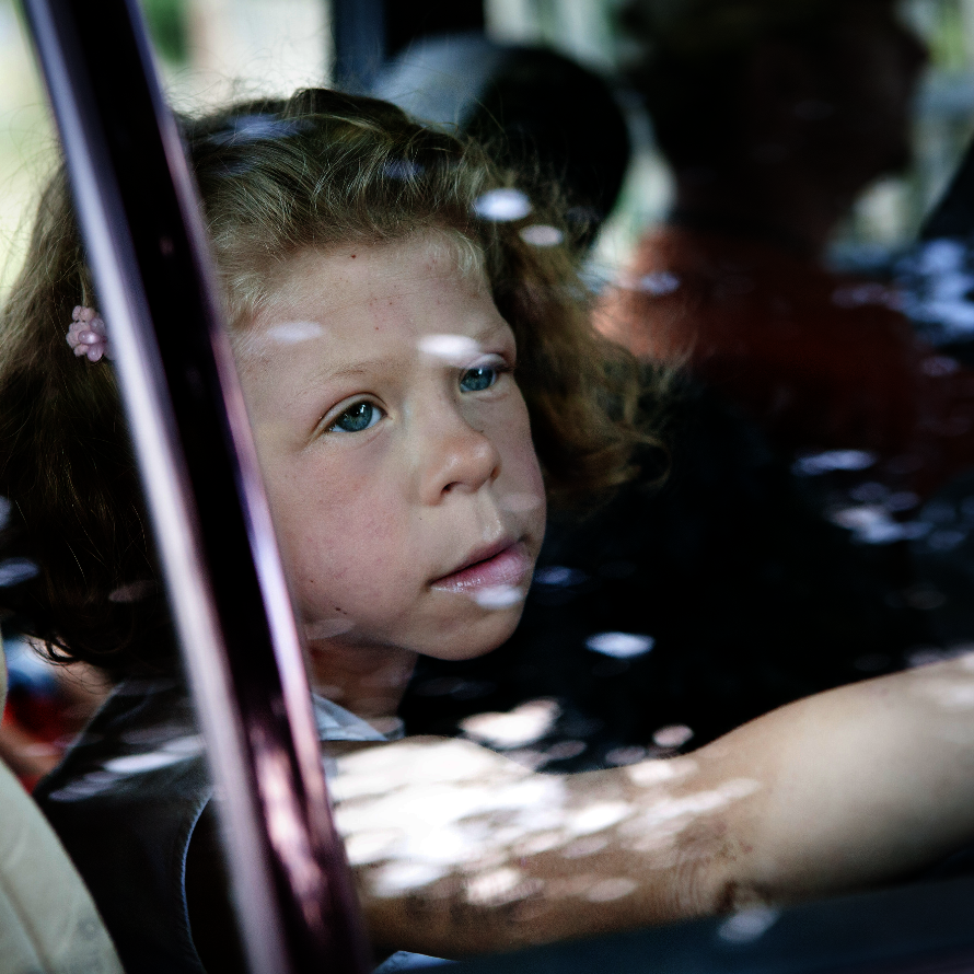 Ein junges Mädchen mit lockigem, blonden Haar sitzt in einem Auto und schaut mit nachdenklichem Blick aus dem Fenster. Ihre Hand ist an die Scheibe gelegt. Die Reflexionen auf dem Glas und die verschwommenen Personen im Hintergrund verleihen dem Bild eine melancholische Stimmung.
