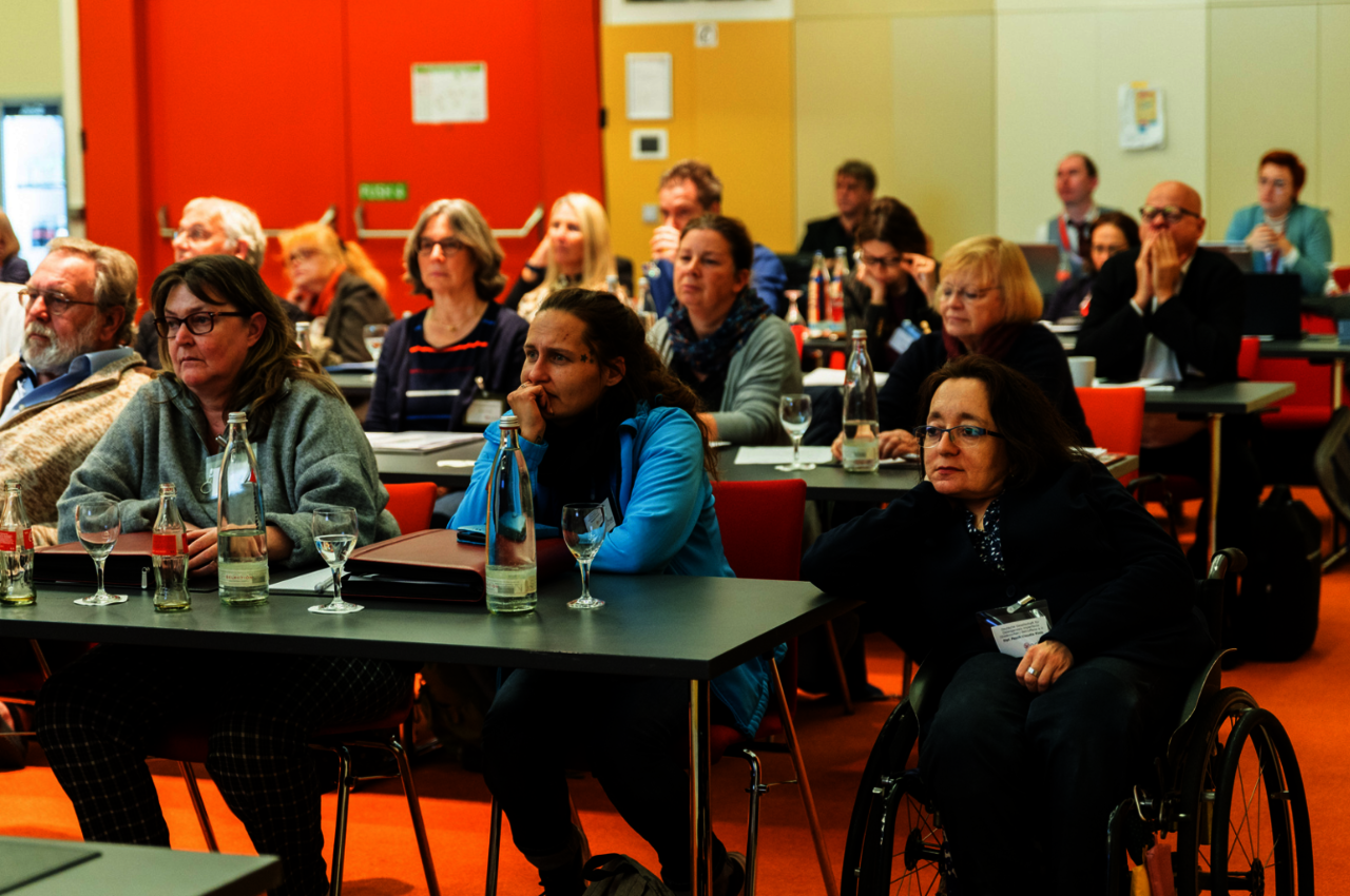 Mitgliederversammlung Eine Gruppe von Menschen sitzt aufmerksam in einem Konferenzraum mit orangefarbenem Teppichboden und einem hellen, modernen Interieur. Die Teilnehmer blicken nach vorne, offenbar einem Vortrag oder einer Präsentation folgend. Vor ihnen stehen Tische mit Getränkeflaschen, Gläsern und Unterlagen. Unter den Anwesenden befindet sich eine Frau im Rollstuhl im Vordergrund, die konzentriert zuhört. Im Hintergrund sind weitere Teilnehmer zu sehen, die eine Vielfalt an Alter und Stil repräsentieren.
