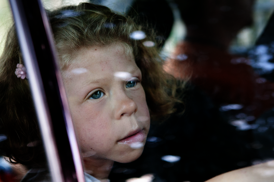 Ein junges Mädchen mit lockigem, blonden Haar sitzt in einem Auto und schaut mit nachdenklichem Blick aus dem Fenster. Ihre Hand ist an die Scheibe gelegt. Die Reflexionen auf dem Glas und die verschwommenen Personen im Hintergrund verleihen dem Bild eine melancholische Stimmung.