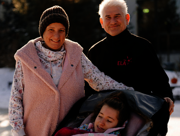 Drei Personen im Freien, eine Person sitzt in einem Kinderwagen mit einer Decke, die anderen beiden stehen daneben, eine trägt eine rosa Weste, die andere ein schwarzes Oberteil mit rotem Schriftzug