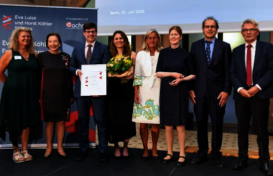 Gruppe von sechs Personen, die zusammenstehen und lächeln. Eine Person hält ein Zertifikat, eine andere einen Blumenstrauß. Alle tragen formelle Kleidung. Im Hintergrund ein Banner mit 'Eva Luise und Horst Köhler Stiftung'.