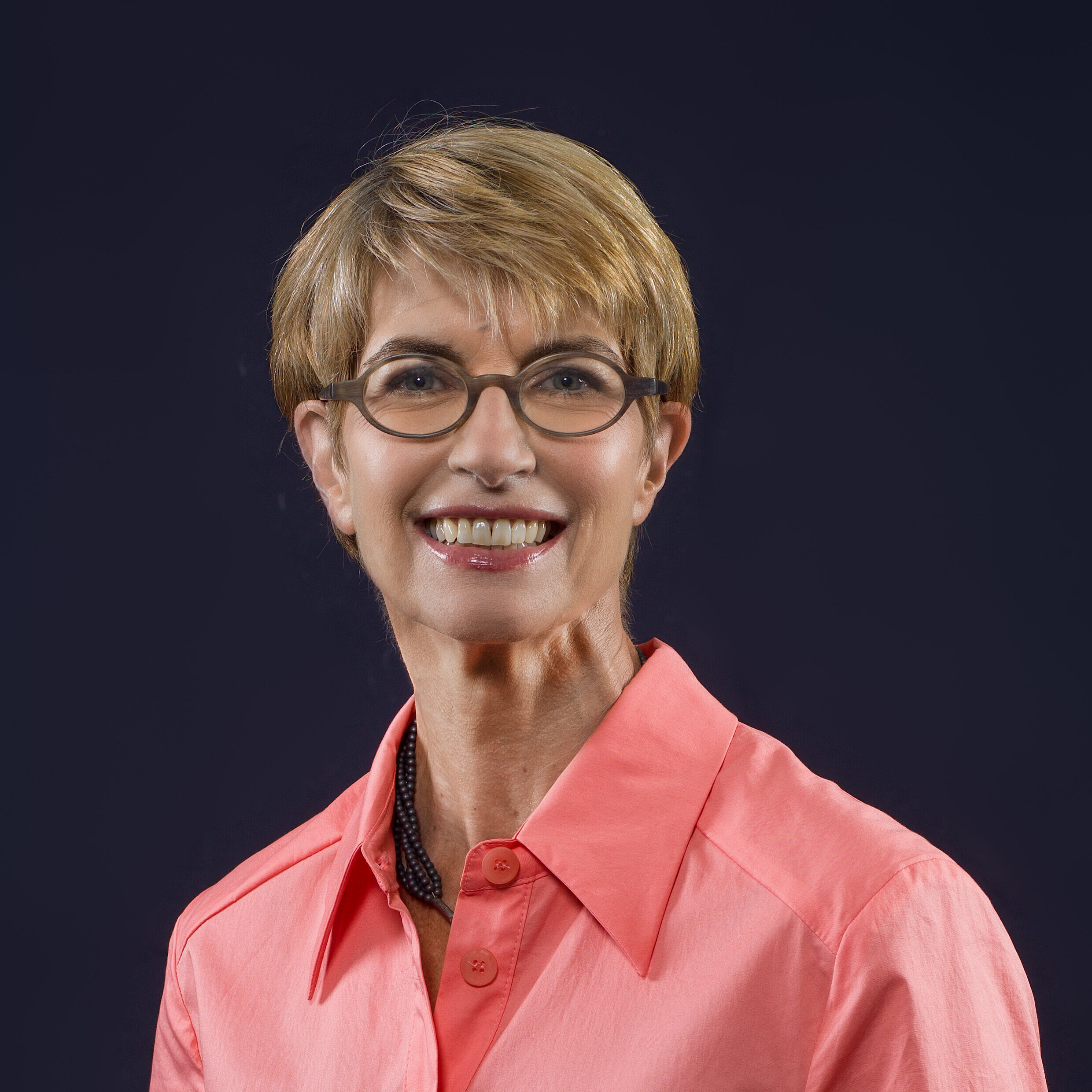 Person mit kurzem blonden Haar trägt ein rotes Hemd mit offenem Kragen vor dunklem Hintergrund.