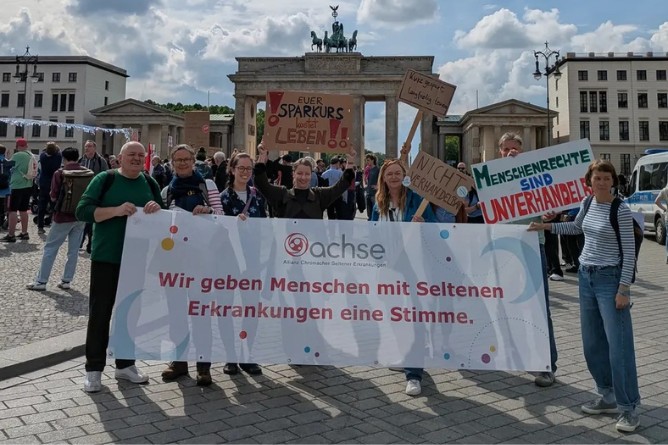 ACHSE Team auf dem Protesttag zur Gleichstellung von Menschen mit Behinderung Gruppe von Menschen vor dem Brandenburger Tor hält Banner mit der Aufschrift 'Wir geben Menschen mit Seltenen Erkrankungen eine Stimme' und weitere Schilder mit Forderungen zu Menschenrechten und Lebenssparplänen