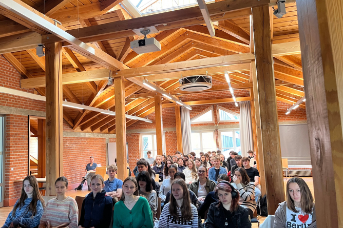 Gruppe von Schülerinnen und Schülern sitzt in einem hellen Raum mit sichtbaren Holzbalken und Backsteinwänden
