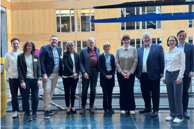 David Gibanica, Jessica Vass, Jannes Moritz, Annette Grüters-Kieslich, Christopher Baum, Christine Mundlos, Nora Seitz, Stephan Albani, Eva Thull, Johannes Heuckeroth Gruppe von neun Personen steht in einem modernen Innenraum vor einer Glasbrüstung und einer Holzpaneelwand