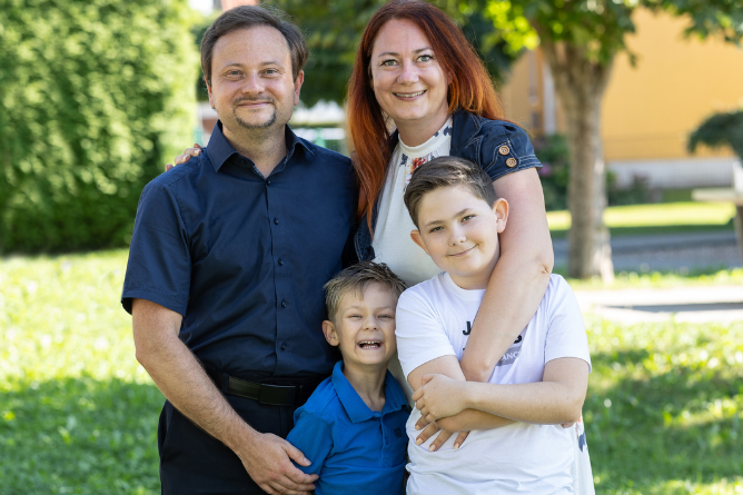 Familie Schneider im Ravensburger Spieleland, © Ravensburger Spieleland Familie mit zwei Kindern steht eng umarmt im Freien vor einem Baum und einem gelben Haus