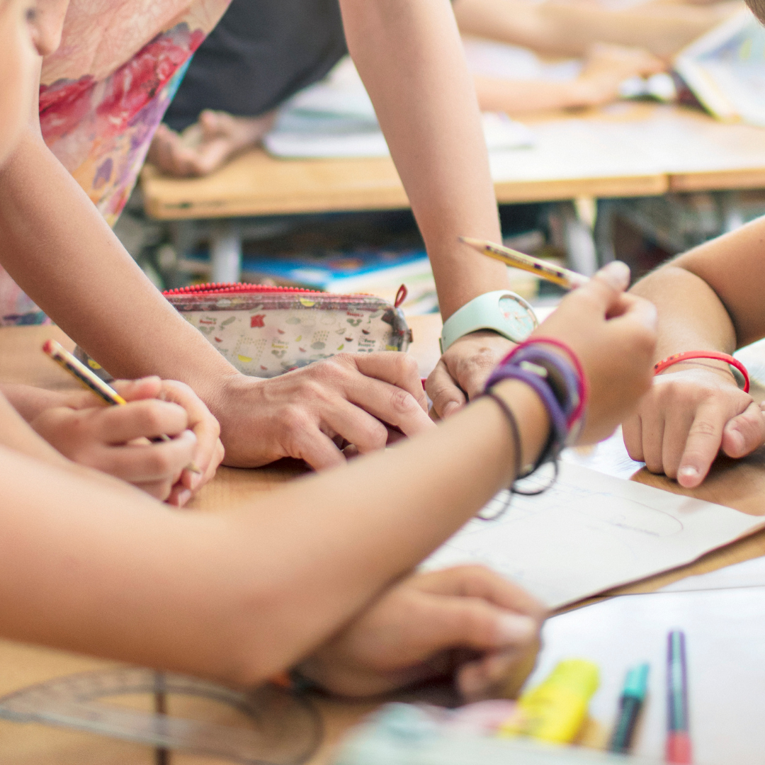 Hände von Kindern und einer erwachsenen Person, die gemeinsam mit einem Bleistift auf einem Tisch schreiben oder zeichnen