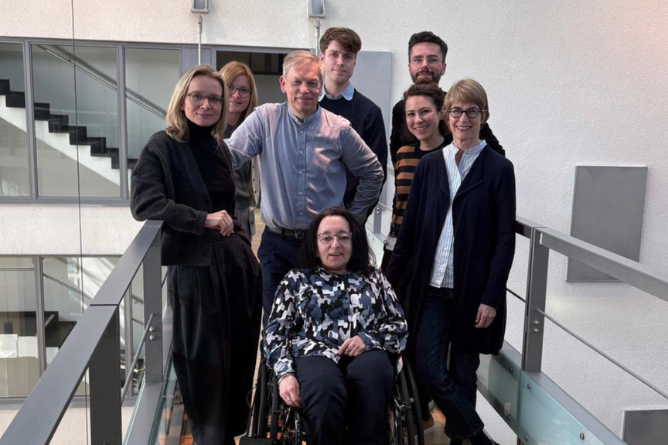 Claudia Müller-Birn (FU), Jelena Maric-Biresev, Lorenz Grigull, Moritz Fischer (Uniklinik Bonn), Peter Sörries (FU), Nadine Scholten (Uniklinik Bonn), Christine Mundlos (ACHSE), Claudia Finis (OI-Gesellschaft), © Nadine Weinstock Gruppe von sechs Personen, darunter eine im Rollstuhl, steht auf einem Balkon mit Glasgeländer in einem modernen Innenraum