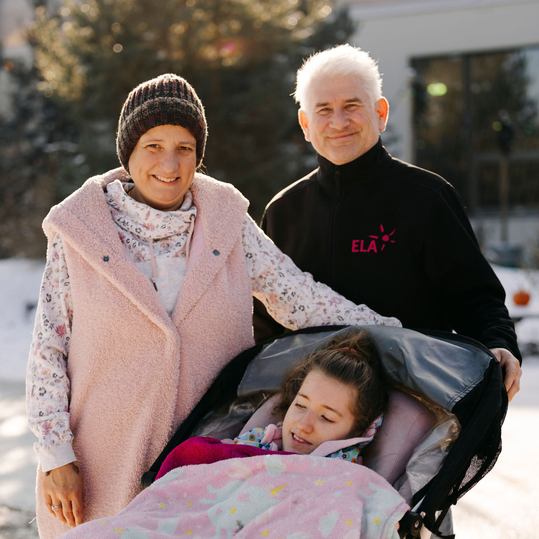 Copyright: Carina Pilz Drei Personen im Freien, eine Person sitzt in einem Kinderwagen mit einer Decke, die anderen beiden stehen daneben, eine trägt eine rosa Weste, die andere ein schwarzes Oberteil mit rotem Schriftzug