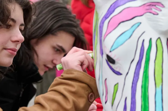 Zebra-Challenge beim Aktionstag im Ravensburger Spieleland Junge Frau mit dunklen Locken und brauner Jacke malt bunte Streifen auf eine weiße Maske