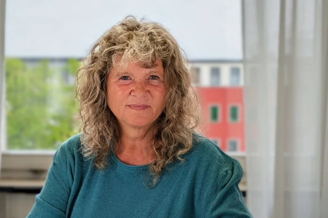 ACHSE Vorsitzende Geske Wehr Frau mit lockigem Haar und grünem Oberteil vor Fenster mit Blick auf grüne Bäume und rotes Gebäude