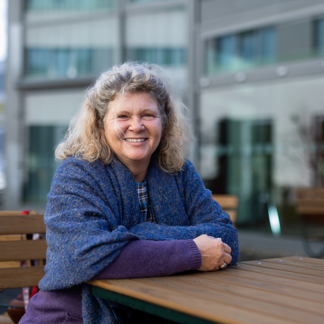 Geske Wehr, Vorstandsvorsitzende ACHSE e.V. Person mit lockigem, schulterlangem Haar sitzt mit verschränkten Armen an einem Holztisch im Freien vor einem modernen Glasgebäude