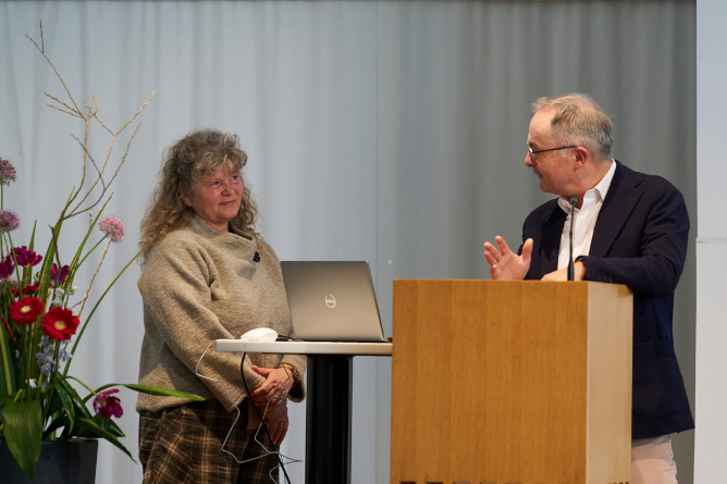 Geske Wehr und Prof. Dr. Martin Wabitsch Zwei Personen stehen an einem Rednerpult und einem kleinen Tisch mit Laptop vor einem hellen Vorhang, links davon ein großer Blumenstrauß