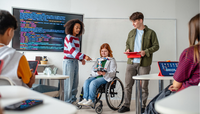 Guter Schulstart braucht sichere Schulbegleitung Drei junge Menschen, eine im Rollstuhl, zeigen in einem Klassenraum mit Computern und großem Bildschirm mit Programmiercode eine technische Konstruktion
