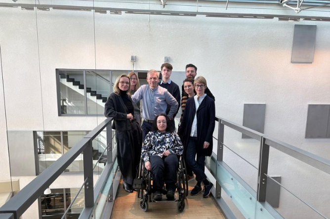 Claudia Müller-Birn (FU), Jelena Maric-Biresev, Lorenz Grigull, Moritz Fischer (Uniklinik Bonn), Peter Sörries (FU), Nadine Scholten (Uniklinik Bonn), Christine Mundlos (ACHSE), Claudia Finis (OI-Gesellschaft), © Nadine Weinstock Gruppe von sechs Personen, darunter eine im Rollstuhl, steht auf einem Balkon mit Glasgeländer in einem modernen Innenraum