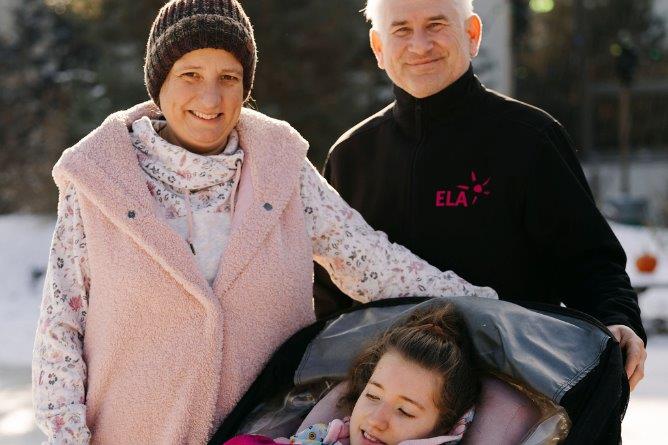 Drei Personen im Freien, eine Person sitzt in einem Kinderwagen mit einer Decke, die anderen beiden stehen daneben, eine trägt eine rosa Weste, die andere ein schwarzes Oberteil mit rotem Schriftzug