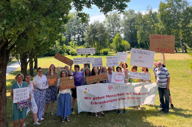 Laut gegen das PNOG Gruppe von Menschen steht im Freien auf einer Wiese und hält Schilder sowie ein Banner mit der Aufschrift 'Wir geben Menschen mit Seltenen Erkrankungen eine Stimme'