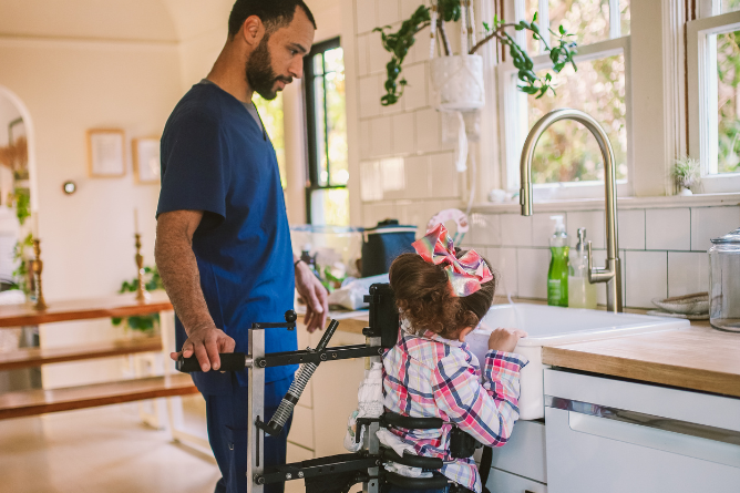 Petition "Stoppt den AKI-Pflegekollaps" Ein Mann in blauer Kleidung steht neben einem Kind im Rollstuhl in einer hellen Küche mit Fenster und Pflanzen