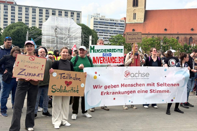 ACHSE auf dem Protesttag zur Gleichstellung von Menschen mit Behinderungen Gruppe von Menschen bei einer Demonstration mit Bannern und Schildern, darunter ein Banner mit dem Text 'Wir geben Menschen mit Seltenen Erkrankungen eine Stimme' und Schildern mit den Aufschriften 'Kürzungen Zukunft', 'Teilhabe Solidarität' und 'Menschenrechte sind unverhandelbar'