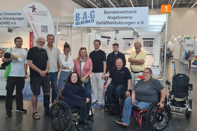Gruppenbild REHACARE 2026 Gruppe von Menschen, darunter mehrere im Rollstuhl, vor einem Messestand mit Bannern des Bundesverbands Angeborene Gefäßerkrankungen e.V.