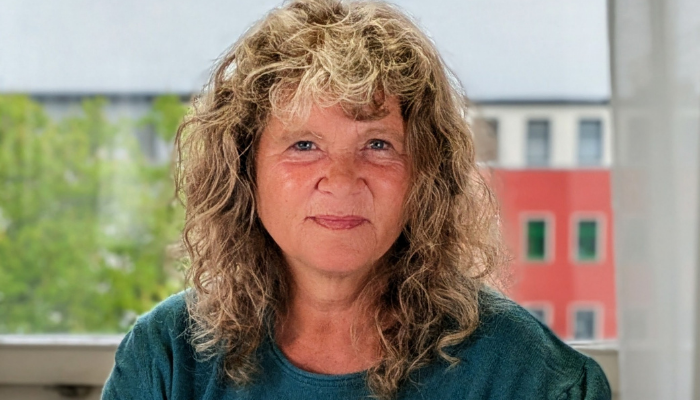Geske Wehr Frau mit lockigem Haar und grünem Oberteil vor Fenster mit Blick auf grüne Bäume und rotes Gebäude