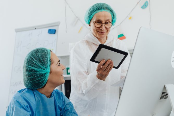 Zwei Personen in medizinischer Schutzkleidung und Haarnetzen vor einem Computer, eine hält ein Tablet