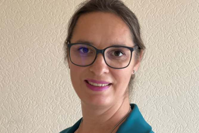 Anika Schlaud Frau mit Brille und zurückgebundenem Haar trägt ein türkisfarbenes Sakko und lächelt vor einer hellen, strukturierten Wand