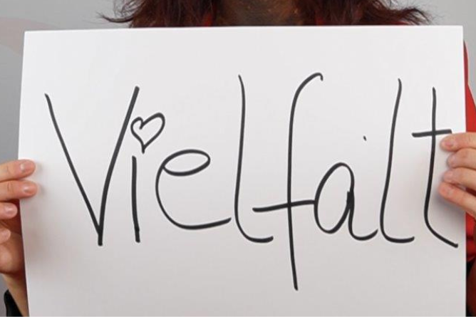 Nanée mit dem Schild: Vielfalt Person hält ein weißes Schild mit dem handgeschriebenen Wort 'Vielfalt' und einem kleinen Herz über dem Buchstaben 'i'