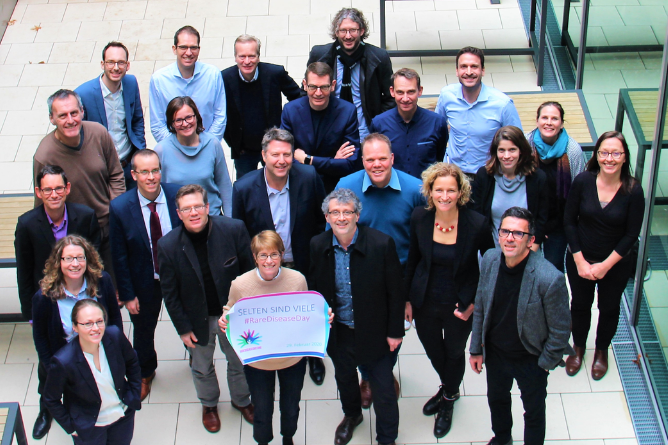 Teilnehmende am Symposium "Translationale Forschung" Gruppe von Menschen in Geschäftskleidung steht in einem Innenraum mit Fliesenboden, eine Person hält ein Schild mit dem Text 'SELTEN SIND VIELE #RareDiseaseDay'