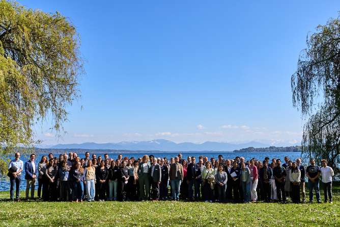 Teilnehmende am Symposium von Research4Rare Große Gruppe von Menschen steht auf einer Wiese vor einem See mit Bergen im Hintergrund und Bäumen an den Seiten