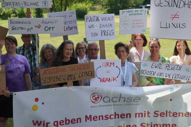 Anika Schlaud (2. v. r.) während der ACHSE Sommerwerkstatt Gruppe von Menschen hält Schilder mit Botschaften zu Armut, Krankheit und Politik sowie ein Banner mit dem Schriftzug 'achse Allianz Chronischer Seltener Erkrankungen'