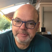 Porträt von Jürgen Grunert, einem Mann mit Brille und Glatze. Er trägt ein dunkles T-Shirt und sitzt in einer entspannten Umgebung. Im Hintergrund sind Lichter und eine Fensterfront zu sehen.