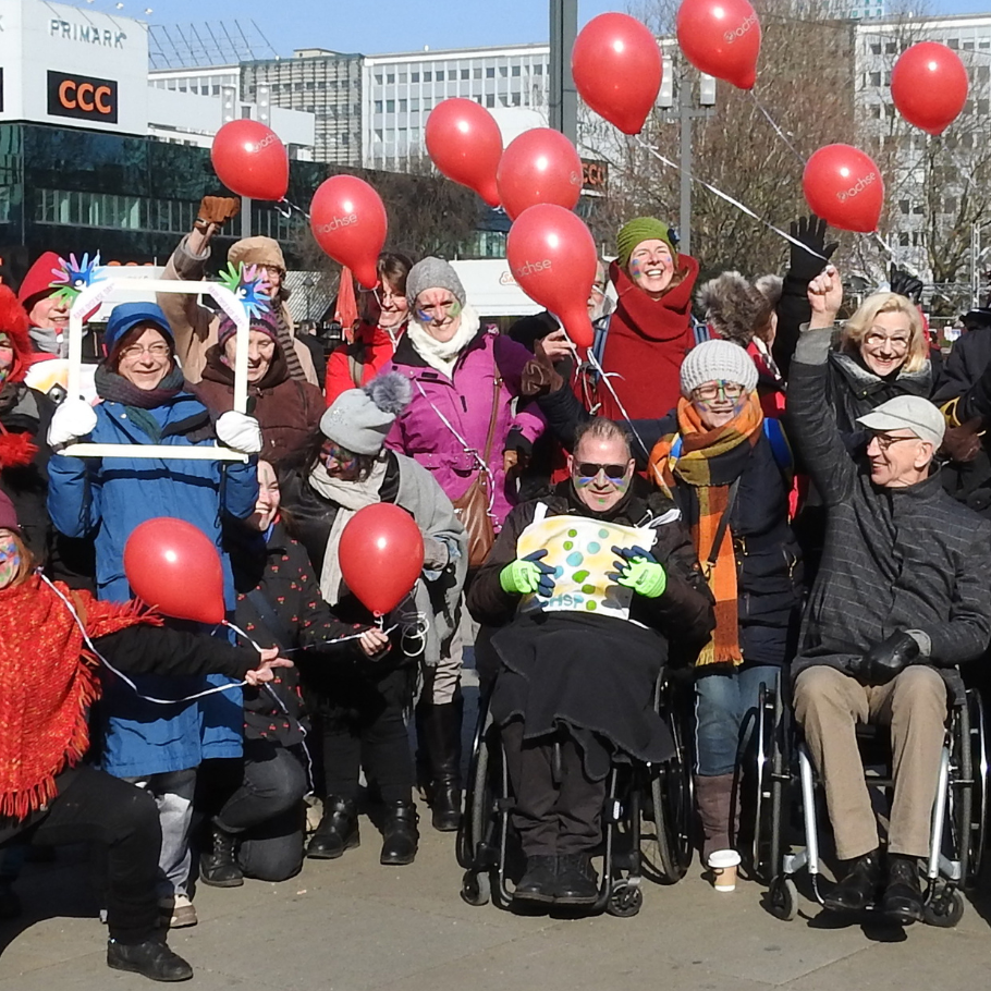 Eine Gruppe von Menschen hat sich bei sonnigem Wetter im Freien versammelt. Einige von ihnen halten rote Luftballons mit dem "ACHSE"-Logo, während andere fröhlich winken oder in die Kamera lächeln. In der Mitte des Bildes sind zwei Personen in Rollstühlen zu sehen. Die Gruppe ist bunt gekleidet, mit Schals, Mützen und Jacken, passend zu der kühlen Witterung. Im Hintergrund sind Gebäude und Geschäfte zu erkennen.