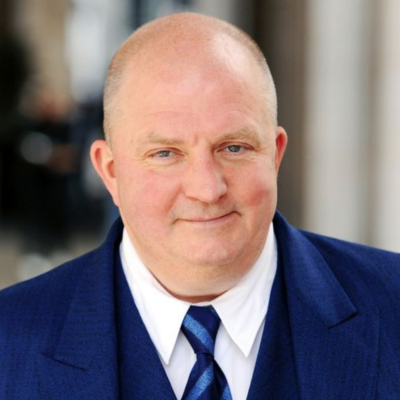 Porträt eines Mannes mit Glatze in einem blauen Anzug, weißem Hemd und einer blauen gestreiften Krawatte. Er lächelt leicht und blickt direkt in die Kamera. Der Hintergrund ist unscharf und deutet auf eine Außenaufnahme hin.