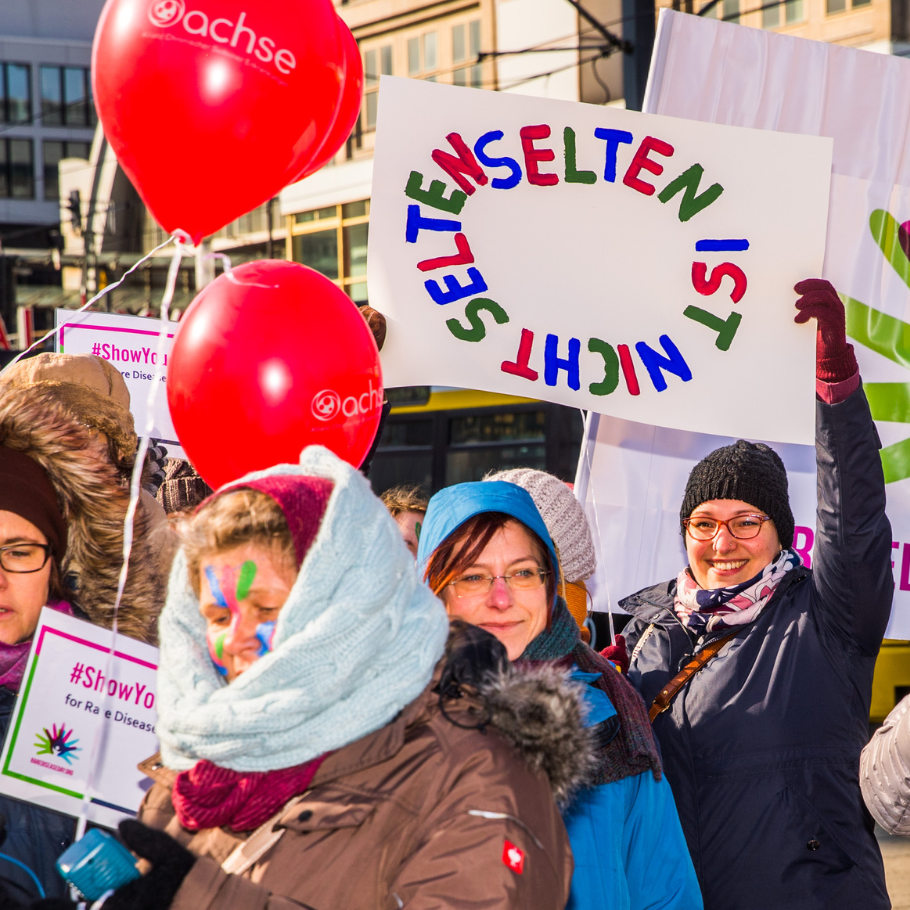 Eine Gruppe von Menschen versammelt sich zum Rare Disease Day im Freien und hält rote Ballons mit dem Logo der ACHSE e. V. sowie Schilder hoch. Eine Frau hält ein Schild mit dem Text 'Selten ist nicht selten' in bunten Buchstaben. Weitere Teilnehmer tragen bunte Gesichtsbemalungen und warme Winterkleidung. Im Hintergrund sind Gebäude und ein Banner des Rare Disease Day zu sehen.