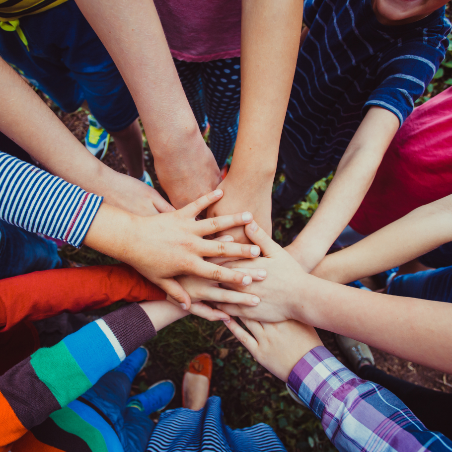 Ein Foto von mehreren Händen, die sich in der Mitte stapeln, als Symbol für Gemeinschaft und Zusammenarbeit. Die Personen tragen bunte Kleidung, darunter gestreifte und karierte Muster. Der Hintergrund zeigt Gras und vereinzelt Schuhe, die zu den stehenden Personen gehören.