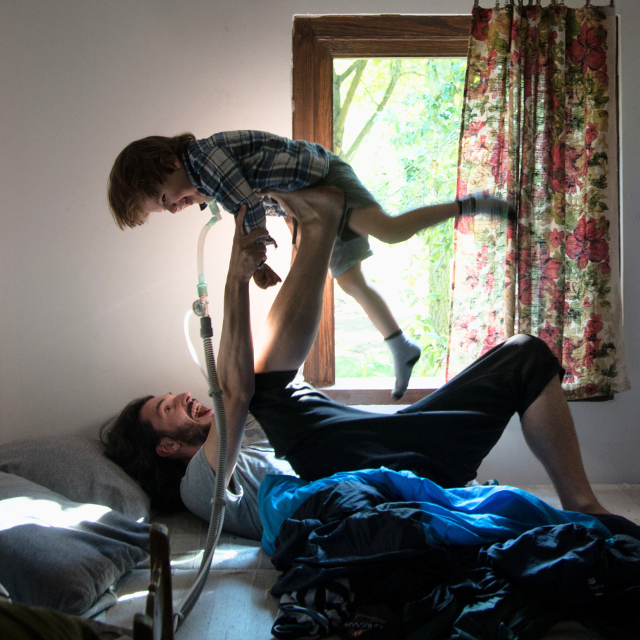 Ein Mann liegt lachend auf einem Bett und hebt ein kleines Kind in die Luft, das ebenfalls lacht. Das Kind hat eine Trachealkanüle am Hals, an die ein Schlauch angeschlossen ist, um ihm Luft zuzuführen. Im Hintergrund ist ein Fenster mit floralem Vorhang zu sehen, durch das Tageslicht und grüne Bäume hineinscheinen.